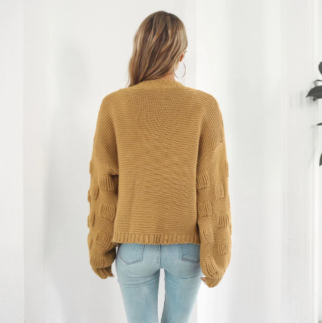 Femme de dos portant un pull en maille beige à manches longues et un jean bleu clair, posant dans un intérieur lumineux. Mode automne-hiver tendance.