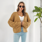 Femme portant un cardigan en tricot beige, lunettes de soleil, top blanc et jean bleu, posant devant une plante verte. Mode automne chic et décontractée.