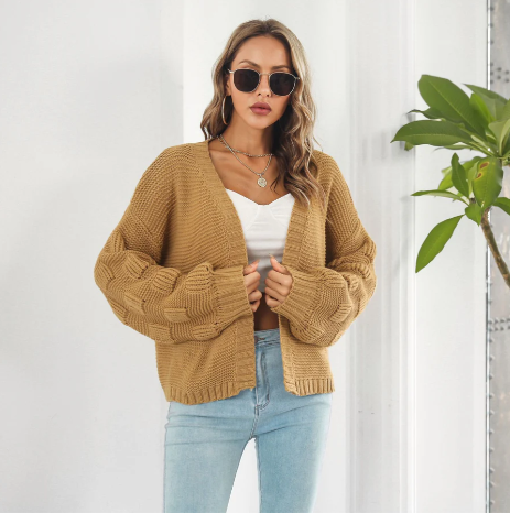Femme portant un cardigan en tricot beige, lunettes de soleil, top blanc et jean bleu, posant devant une plante verte. Mode automne chic et décontractée.