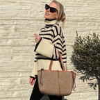 Femme portant un pull rayé noir et blanc, lunettes de soleil, et un sac à main beige en feutre, posant devant un mur en pierre.