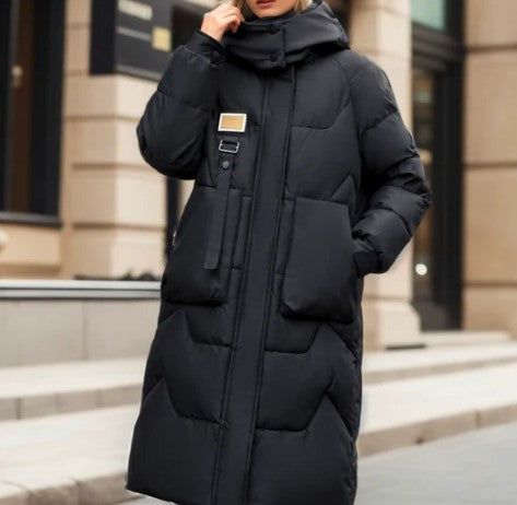 Femme portant un manteau long noir matelassé avec capuche, devant un bâtiment moderne. Mode hiver, vêtement chaud, style urbain élégant.