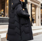 Manteau long noir matelassé avec capuche, porté par une femme en ville. Mode hiver élégante, tendance et confortable pour temps froid.