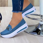 Chaussures mocassins bleues pour femme avec semelles blanches épaisses, ornées d'un détail métallique doré en forme de H, portées avec un jean.