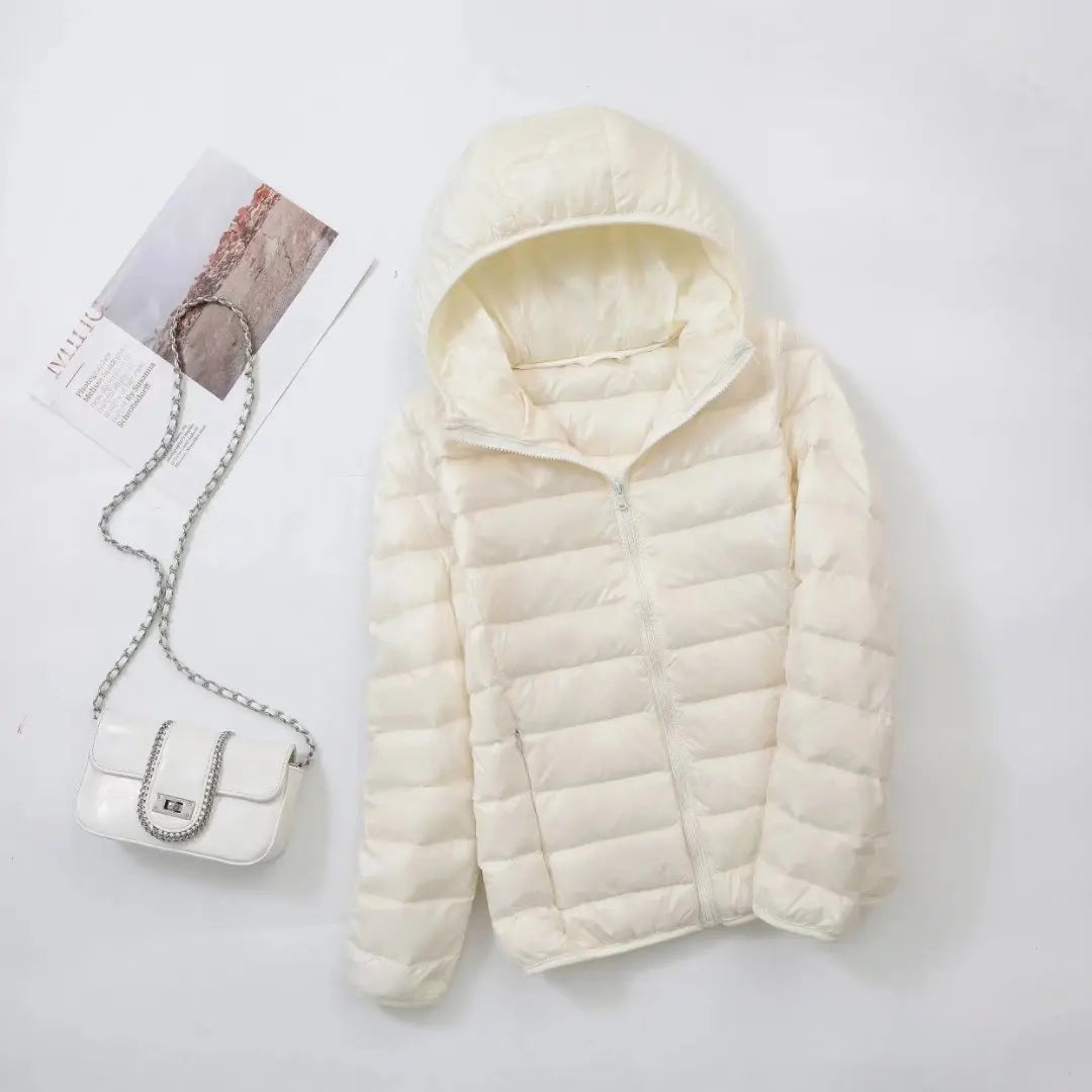 Veste matelassée blanche avec capuche, posée à côté d'un sac à main blanc à chaîne argentée et d'un magazine sur fond blanc. Mode hiver élégante.