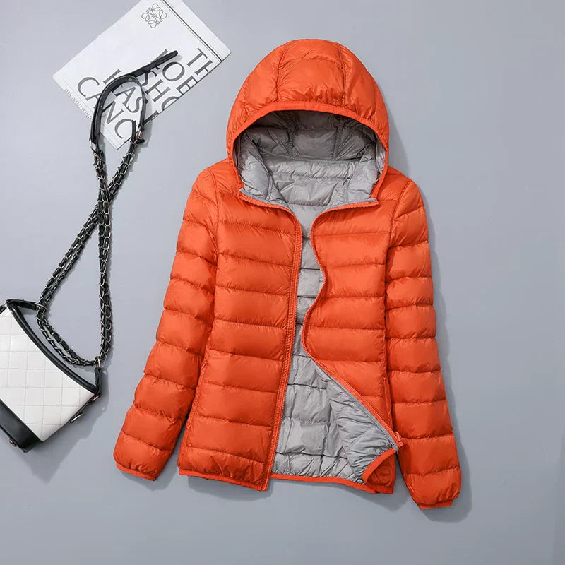 Doudoune orange à capuche, doublure grise, posée à plat sur fond gris avec sac à main blanc et noir. Mode hiver, veste chaude, tendance.