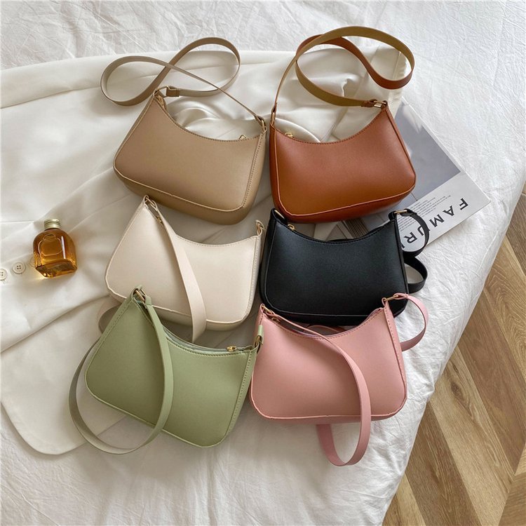 Six sacs à main en cuir de différentes couleurs (beige, marron, blanc, noir, vert, rose) posés sur un lit blanc, avec une bouteille de parfum.