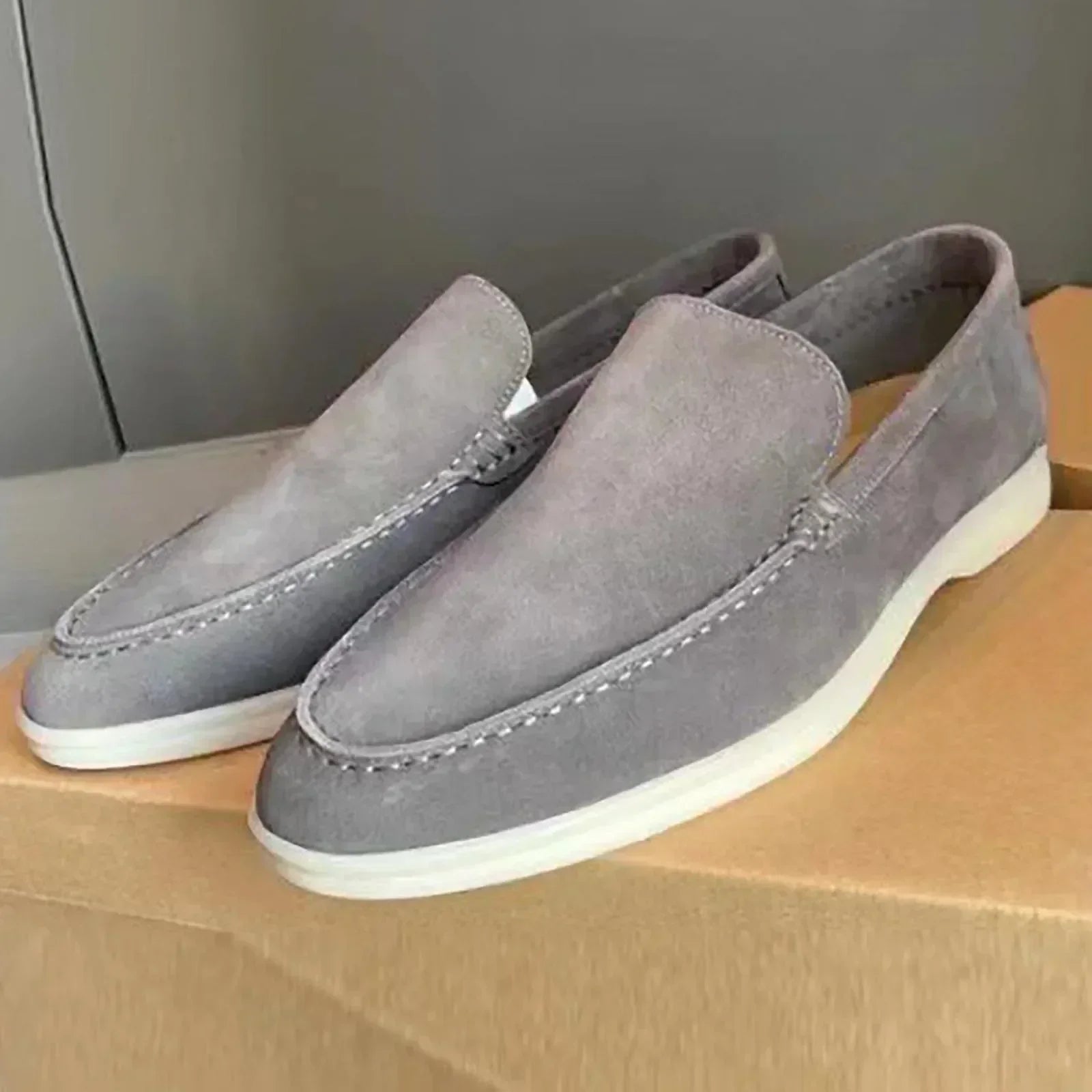 Mocassins en daim gris pour homme sur boîte en carton, style décontracté et élégant, semelle blanche, chaussures tendance et confortables.