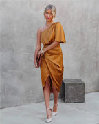 Femme en robe asymétrique dorée, tenant une pochette marron, posant devant un mur gris. Mode élégante, tenue de soirée chic, style moderne.