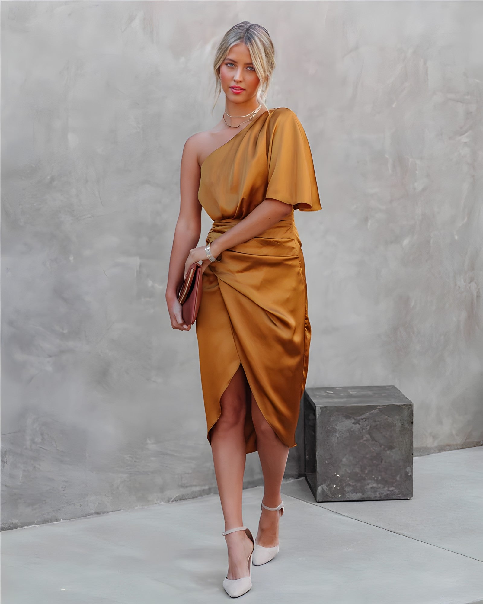 Femme en robe asymétrique dorée, tenant une pochette marron, posant devant un mur gris. Mode élégante, tenue de soirée chic, style moderne.