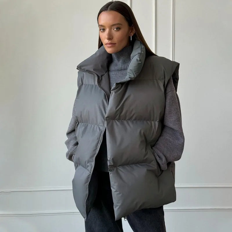 Femme portant une doudoune grise sans manches, col montant, sur un pull gris, posant devant un mur blanc. Mode hiver, style décontracté.