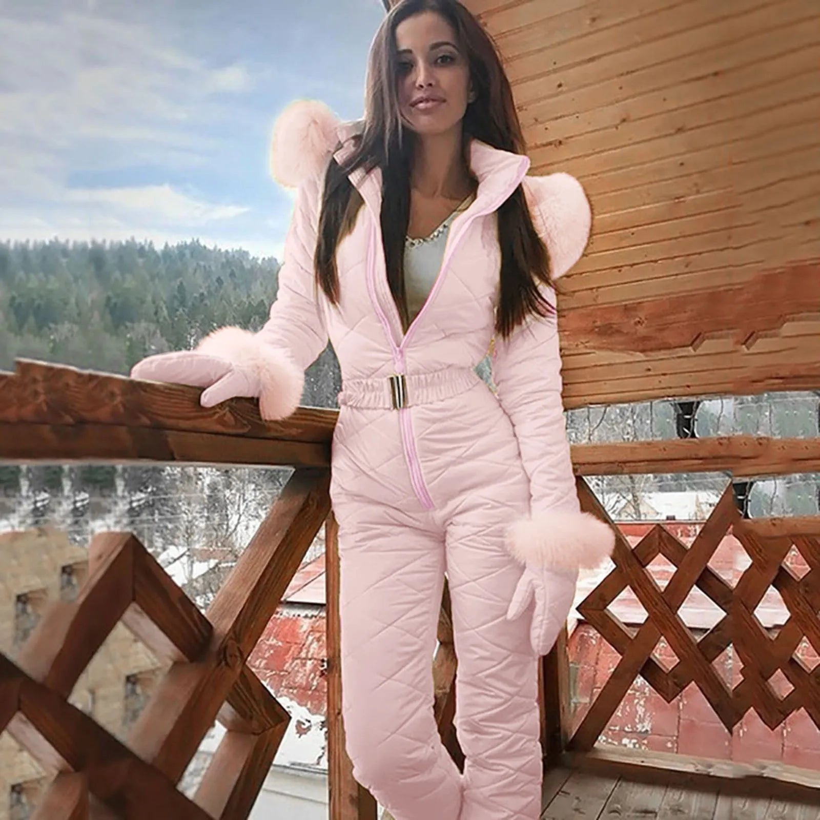 Femme en combinaison de ski rose avec fourrure, posant sur un balcon en bois avec vue sur forêt enneigée. Mode hiver, tenue de ski élégante.