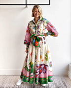 Robe longue colorée avec motifs floraux et animaux, manches bouffantes, ceinture assortie, femme souriante, intérieur moderne, sol en bois.