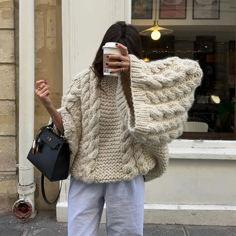 Femme portant un pull en laine torsadée beige, tenant un café à emporter, sac à main noir, devant une vitrine parisienne. Mode automne chic.