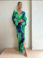 Femme portant une robe longue plissée verte et bleue, posant contre un mur texturé. Mode élégante, tenue de soirée chic, style moderne.