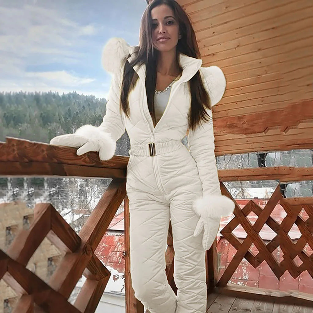 Femme en combinaison de ski blanche avec fourrure, posant sur un balcon en bois, paysage enneigé en arrière-plan, style hiver élégant.