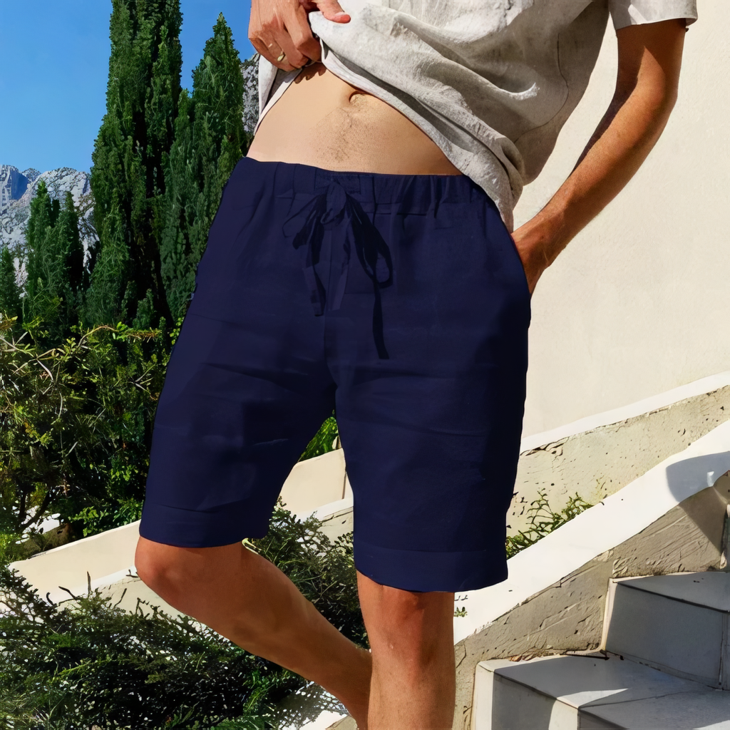 Homme portant un short bleu marine, debout sur des escaliers extérieurs ensoleillés, avec des arbres et montagnes en arrière-plan. Mode estivale décontractée.