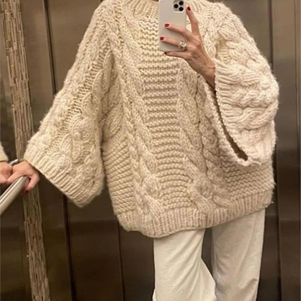 Femme portant un pull en laine torsadé beige avec manches larges, tenant un smartphone dans un ascenseur. Mode automne-hiver, style décontracté.