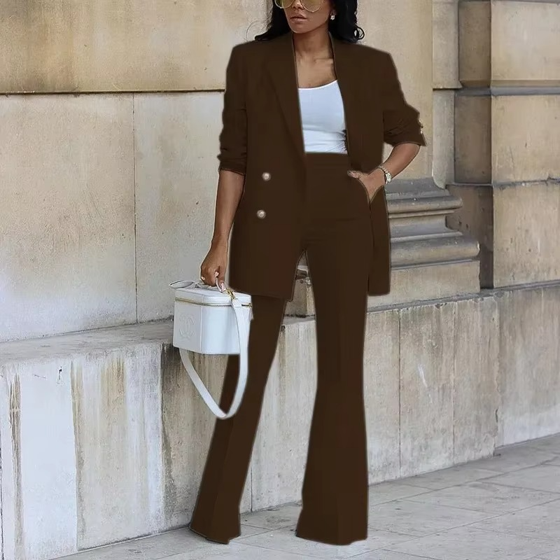 Femme élégante en tailleur marron, top blanc, tenant un sac à main blanc, posant devant un mur en pierre. Mode chic, style urbain, tendance.