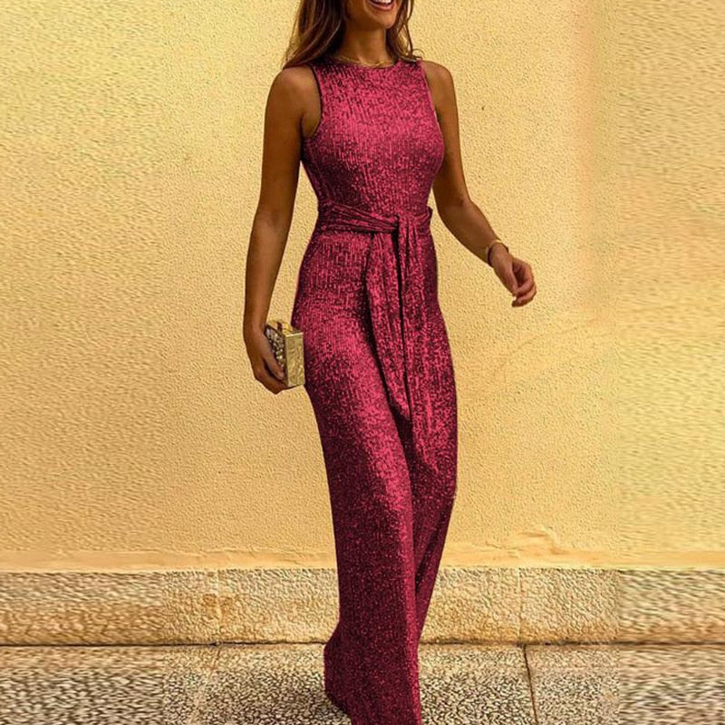 Robe longue pailletée rose, sans manches, avec ceinture, portée par une femme souriante tenant une pochette dorée. Mode élégante soirée.