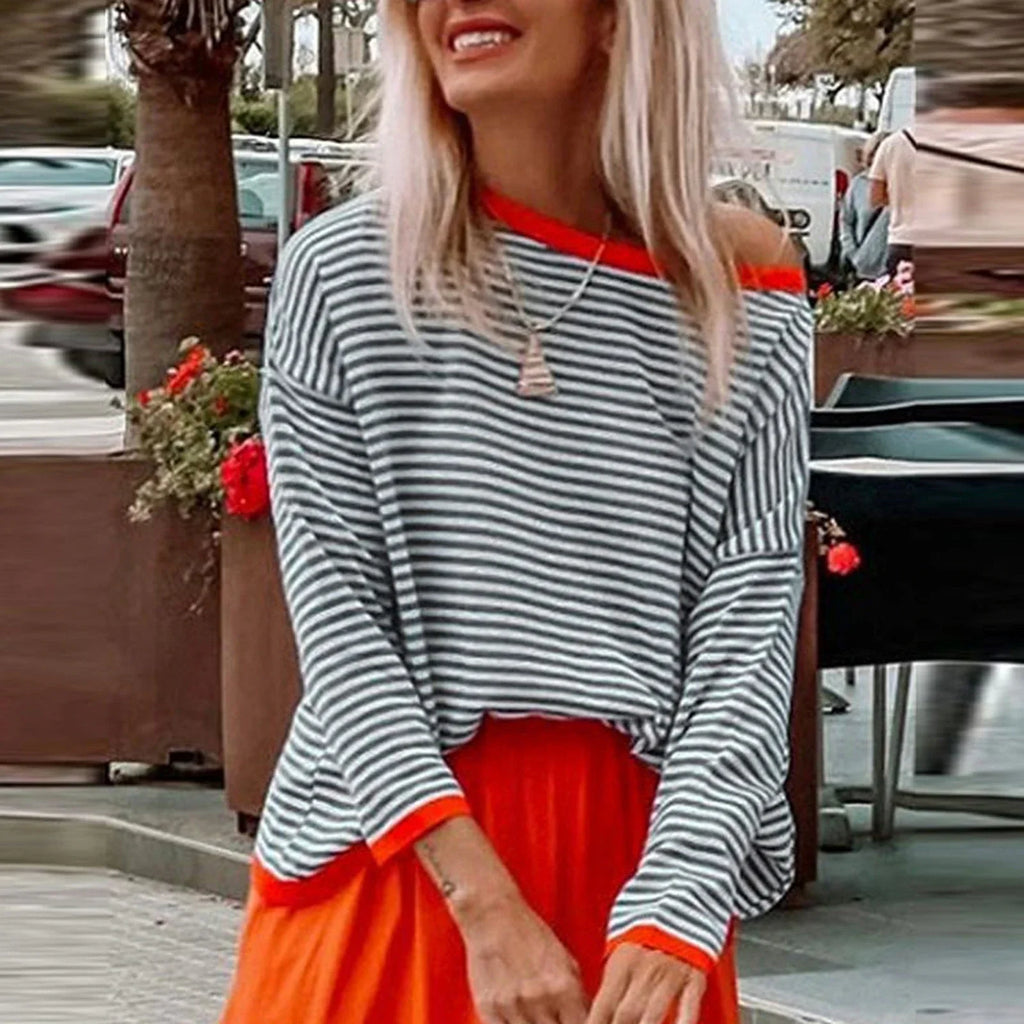 Femme souriante en pull rayé noir et blanc, jupe orange vif, collier pendentif, posant en extérieur urbain avec plantes en arrière-plan.