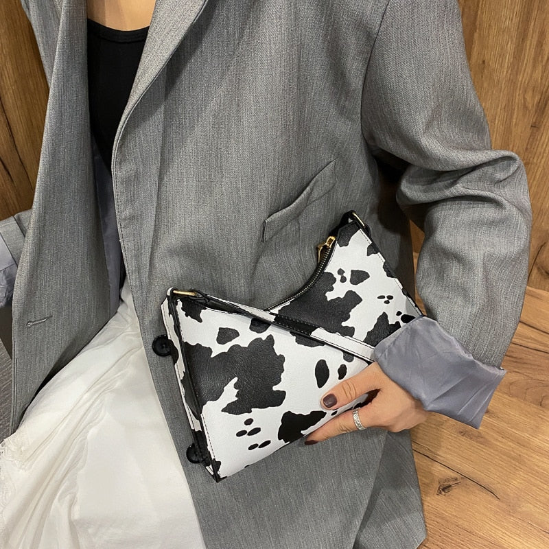 Sac à main motif vache noir et blanc, tenu par une personne en blazer gris et jupe blanche, posé sur un fond en bois. Accessoire mode tendance.