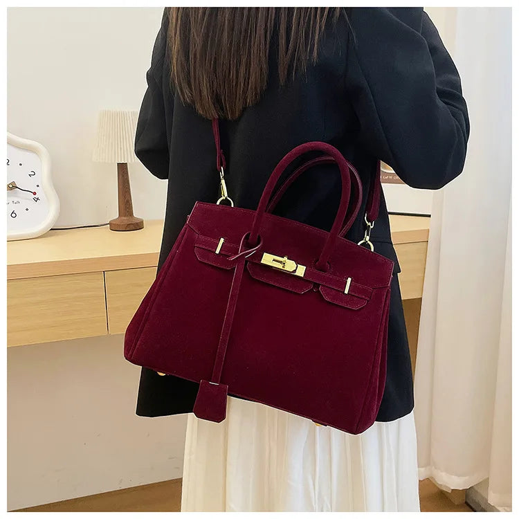 Sac à main en velours bordeaux porté par une femme en manteau noir, devant une table en bois avec une horloge blanche et une lampe. Accessoire élégant.