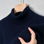 Pull bleu marine en cachemire avec col roulé, suspendu sur un cintre en bois. Main touchant le tissu doux. Mode hiver, vêtement chaud et élégant.