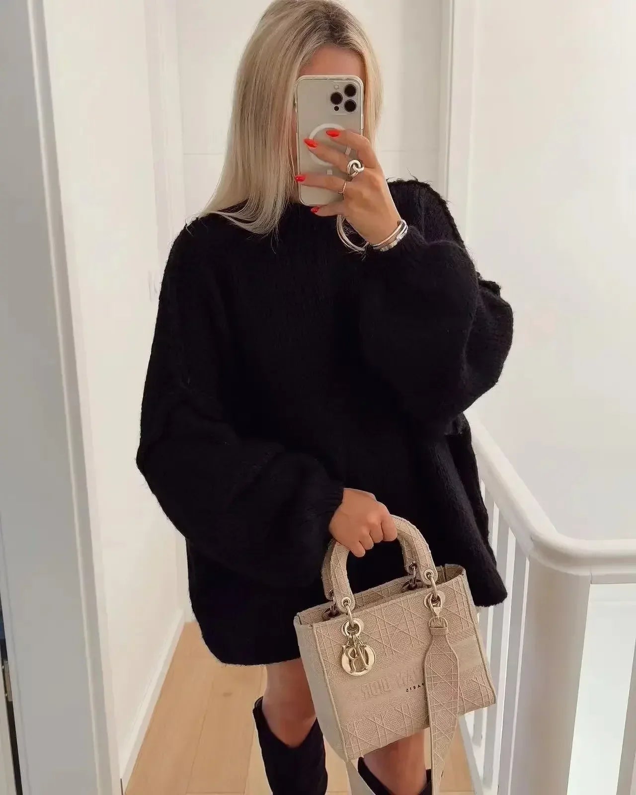 Femme en pull noir oversize, tenant un sac à main beige élégant, prenant un selfie avec un smartphone. Mode automne, style décontracté chic.