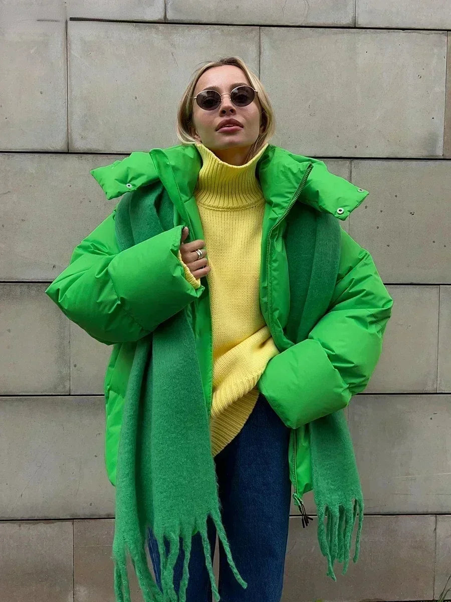 Femme portant une doudoune verte, pull jaune, écharpe verte et lunettes de soleil, posant devant un mur en béton. Mode hiver tendance.