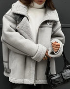 Femme portant un manteau en peau lainée gris avec col en fourrure, pull blanc à col roulé, sac à main noir. Mode hiver élégante et tendance.