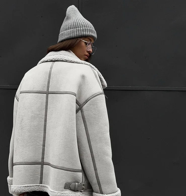 Personne portant une veste en laine grise avec col en fourrure et bonnet assorti, posant devant un mur noir. Mode hiver, style urbain.