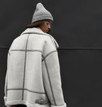 Personne portant une veste en laine grise avec col en fourrure et bonnet assorti, posant devant un mur noir. Mode hiver, style urbain.