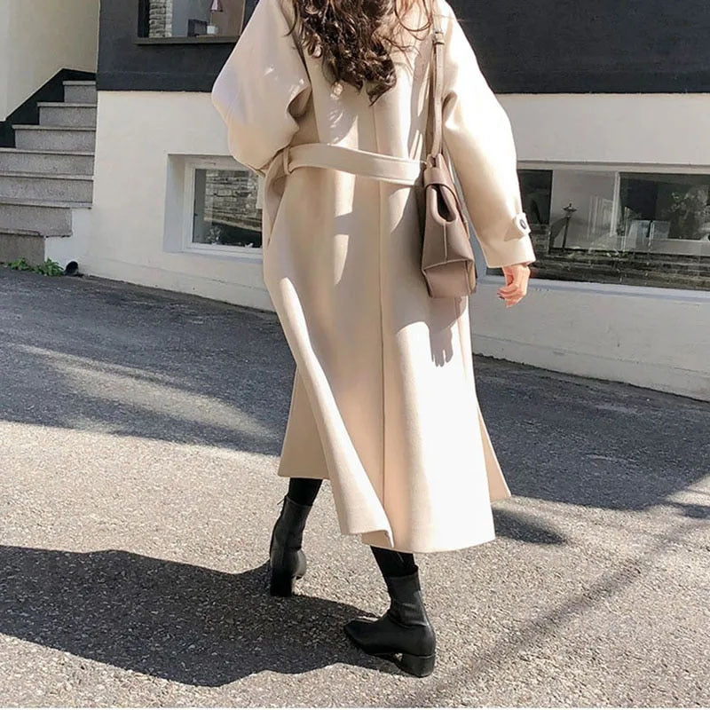 Femme portant un manteau beige élégant, sac à main marron, bottes noires, marchant en extérieur sur un trottoir ensoleillé. Mode automne chic.