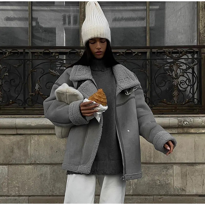 Jeune femme en manteau gris et bonnet blanc tenant un croissant, debout devant une façade urbaine. Mode hivernale, style décontracté.