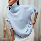 Femme portant un pull bleu clair à col roulé, manches retroussées, posant élégamment. Mode automne-hiver, style décontracté chic.