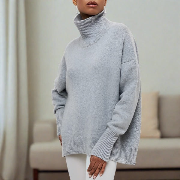 Femme portant un pull gris à col roulé, manches longues, dans un salon moderne. Mode automne-hiver, style décontracté et élégant.