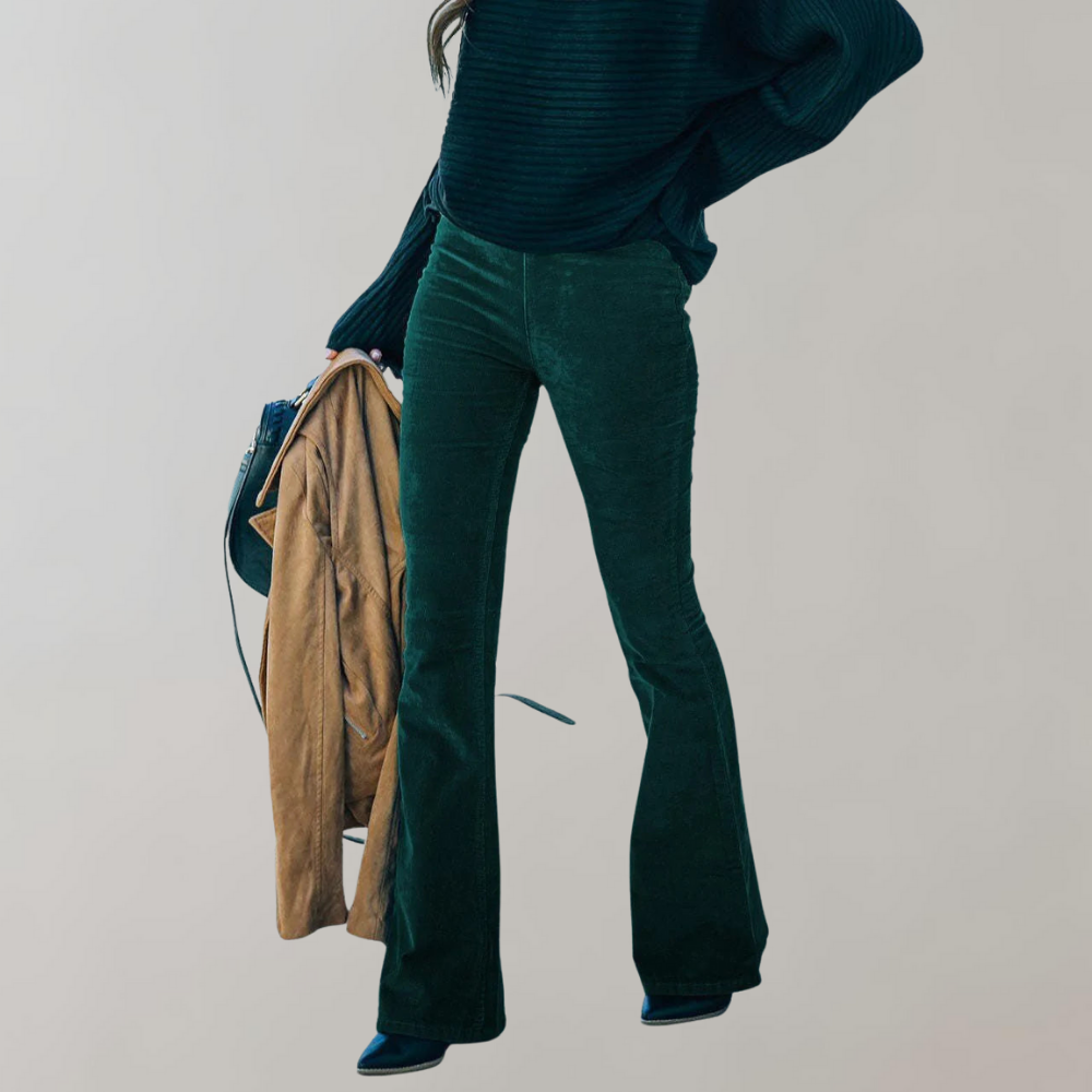Femme en pantalon vert évasé, pull noir, tenant un manteau camel et un sac à main. Mode automne-hiver, style chic et décontracté.