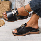 Sandales noires en cuir avec boucle dorée, portées avec un jean et un bracelet turquoise, posées sur un sol en bois clair. Mode estivale élégante.