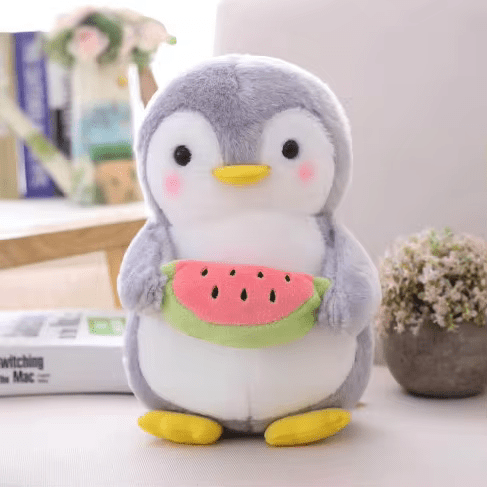 Peluche pingouin mignon tenant une pastèque, posée sur un canapé. Décor intérieur avec livres et plante. Jouet doux pour enfants.
