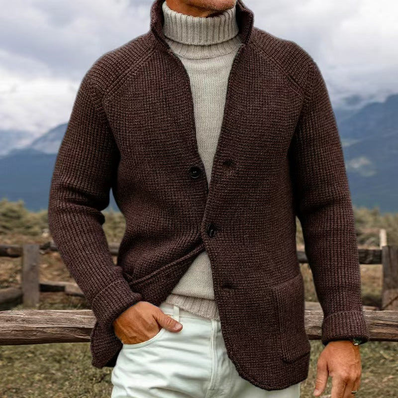 Homme portant un cardigan marron en laine, col roulé beige, debout en extérieur avec montagnes en arrière-plan. Mode automne-hiver élégante.