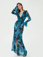 Robe longue transparente bleue à motifs floraux, manches longues évasées, ceinture nouée, portée par une femme posant sur fond blanc.