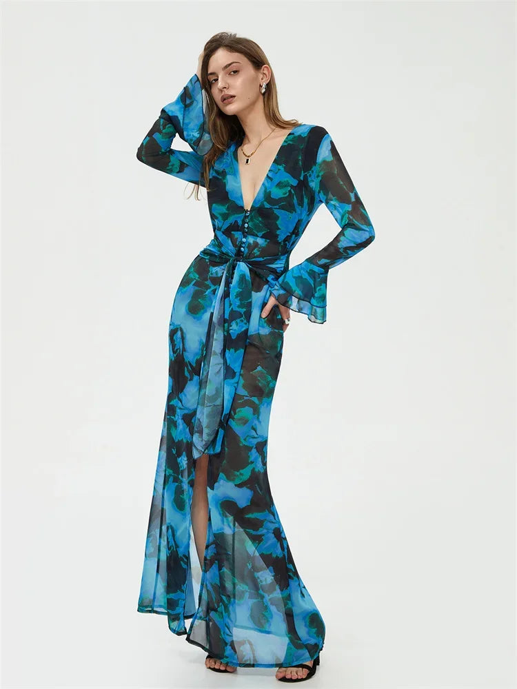 Robe longue transparente bleue à motifs floraux, manches longues évasées, ceinture nouée, portée par une femme posant sur fond blanc.