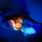 Enfant souriant allongé dans un lit, éclairé par une veilleuse en forme de livre lumineux bleu, créant une ambiance apaisante et magique.