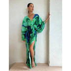 Robe longue verte et bleue à motifs abstraits, manches bouffantes, fente haute, portée par une femme posant contre un mur blanc.