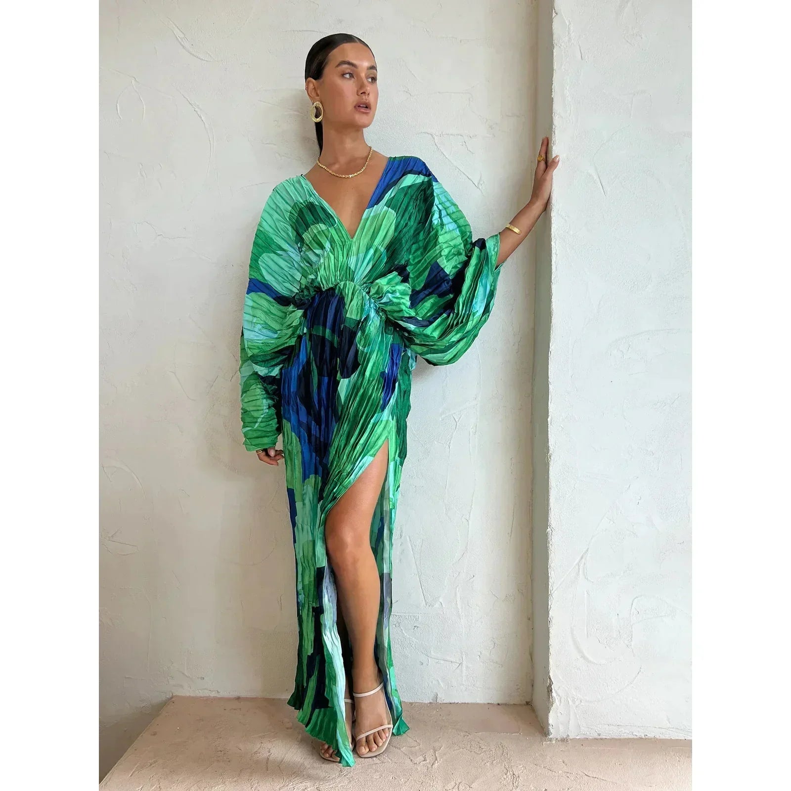 Robe longue verte et bleue à motifs abstraits, manches bouffantes, fente haute, portée par une femme posant contre un mur blanc.