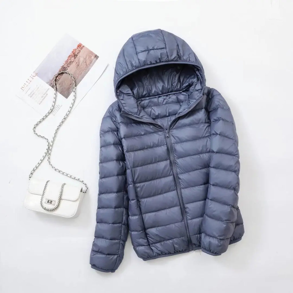 Doudoune bleue à capuche sur fond blanc, accompagnée d'un sac à main blanc et d'une carte postale. Mode hiver, vêtements chauds, accessoires élégants.