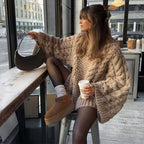 Femme en pull oversize tricoté beige, assise dans un café, tenant un sac à main noir et un gobelet de café, ambiance automnale urbaine.