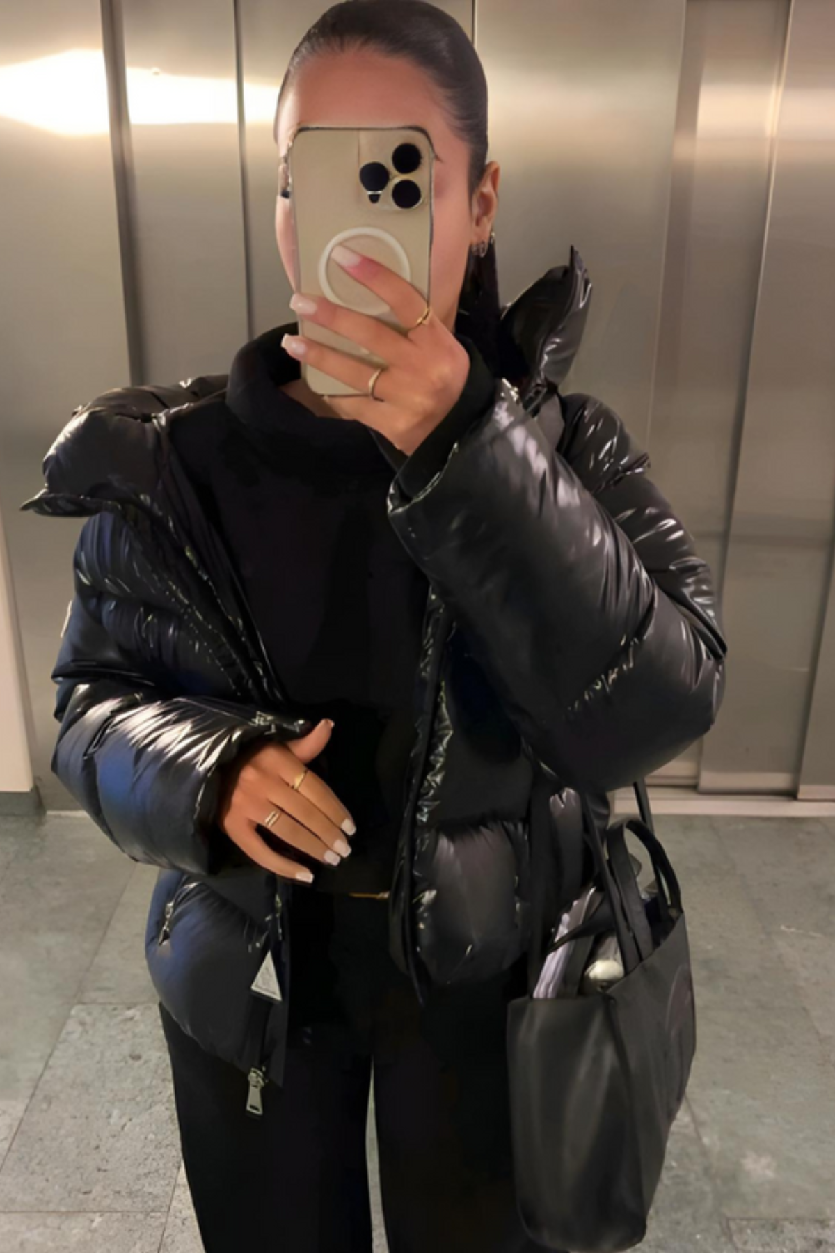 Femme en doudoune noire brillante prenant un selfie dans un ascenseur, tenant un smartphone et un sac noir. Mode hiver, style urbain.