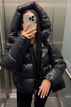 Femme en doudoune noire brillante prenant un selfie dans un ascenseur, capuche relevée, smartphone à la main, style urbain moderne.
