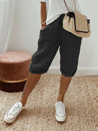 Femme en bermuda noir et t-shirt blanc, portant un sac en osier, debout sur un tapis beige avec des baskets blanches. Mode décontractée chic.